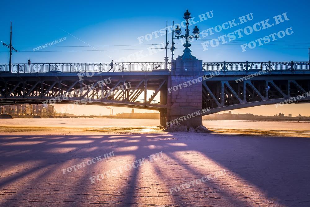 На фотографии изображен Троицкий мост в Санкт-Петербурге зимой на фоне закатного неба. Видны длинные тени от фонарей и замерзшая поверхность реки. Фото может использоваться для туристических буклетов, открыток и рекламных материалов.